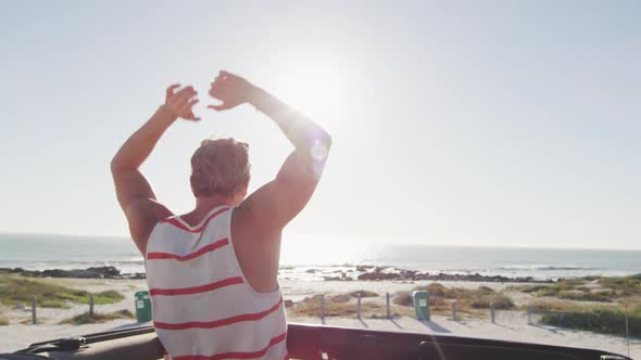 Happy caucasian man standing in convertible car, raising arms and looking out to sea on sunny day