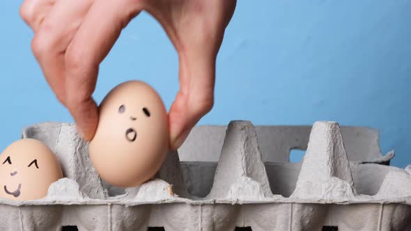 Hand Folds Eggs with Smileys in an Egg Tray on Blue Background