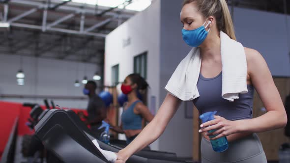 Fit caucasian woman wearing face mask cleaning treadmill machine with disinfectant in the gym