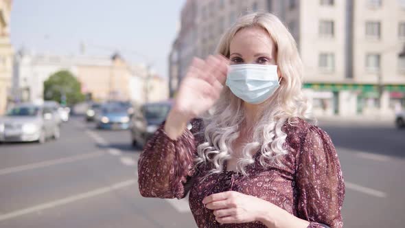 A Middleaged Caucasian Woman in a Face Mask Waves at the Camera with Her Hand in an Urban Area