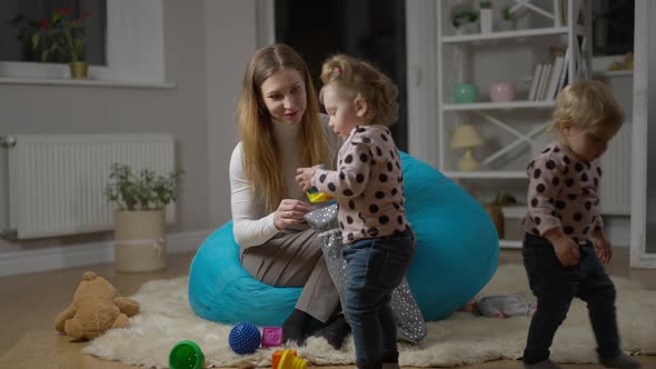 Rack Focus From Smiling Young Millennial Mother to Cute Baby Girl Playing with Toy As Twin Sister