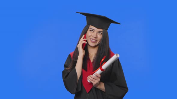 Portrait of Female Student in Graduation Costume Holding Diploma and Talking on a Smartphone