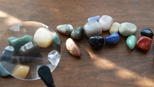 Multicolored semi-precious stones on a wooden table are being explored with a magnifying glass