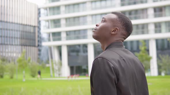 A Young Black Man Looks Around - Office Buildings in the Blurry Background