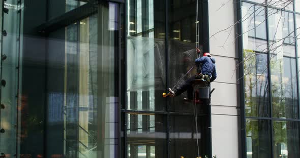 A Man Hanging From Cables Washes the Windows From the Outside of Building