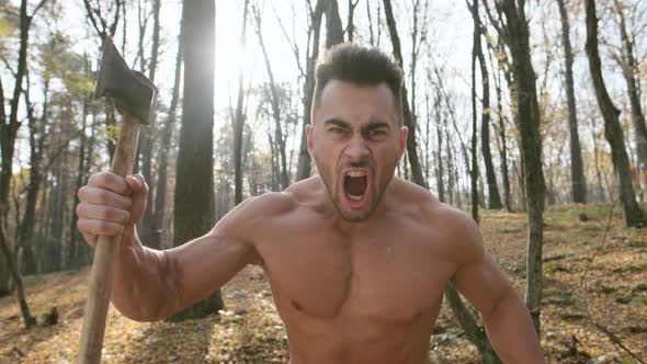 Strong Guy With Naked Torso Holds An Ax In His Hands. Guy In The Woods. He Yells Loudly. Shows Its