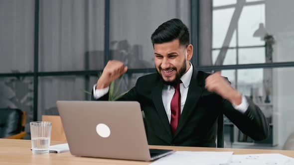 Excited Indian or Arabian Businessman in Suit Sits in a Modern Office Looking at Laptop Happy By