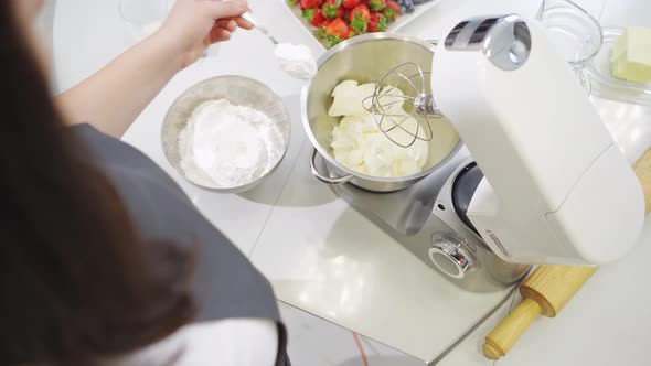 A Young Woman Cook Adds a Spoonful of Flour to Prepare Dough in a Mixer Bowl