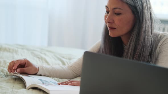 Concentrated Asian woman reading book and looking at laptop