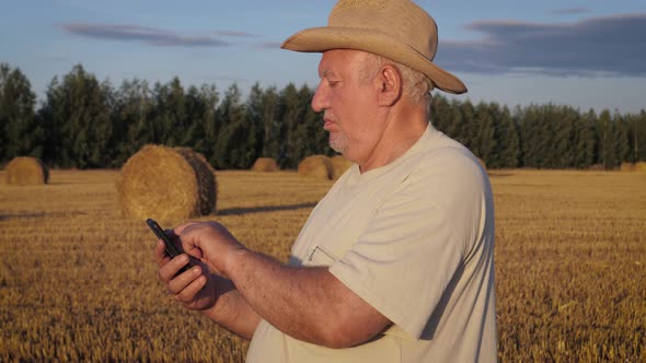 Farmer In Field With Haystacks Uses Smartphone To Control Crop Or Harvesting