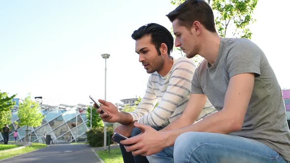 two young multi ethnic men outdoor sitting using smart phone having conversation