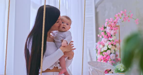 Beautiful Woman Holds a Small Child in Her Arms and Sways with Him on a Swing in a Cozy Light Room