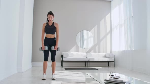 A Young Athletic Woman in a Bright Apartment Performs a Deadlift with Dumbbells at Home Making Leans