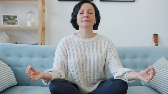 Mature Lady Sitting on Couch in Lotus Pose and Meditating with Eyes Closed Indoors at Home