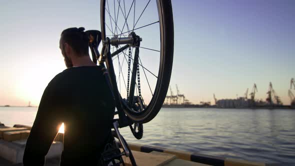 Stylish Handsome Man Walking with Bicycle on His Shoulder During Sunset or Sunrise with Sea Port on