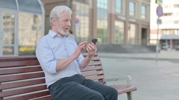 Old Man Celebrating Online Success on Smartphone While Sitting Outdoor on Bench