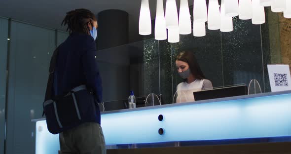 Mixed race man and female receptionist wearing face masks at hotel reception desk