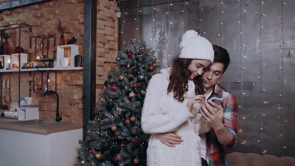 Young Couple Embracing at Home Near the Christmas Tree, Using Their Mobile Phone To Track the New