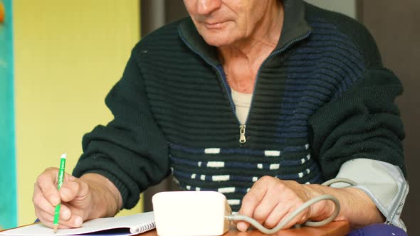 An elderly Caucasian man over 70 at home sitting at a table measures and records the result of the m