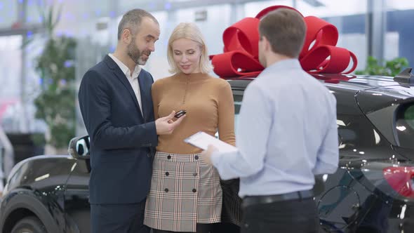 Loving Caucasian Man Giving Car Key To Smiling Beautiful Woman in Showroom with Blurred Dealer