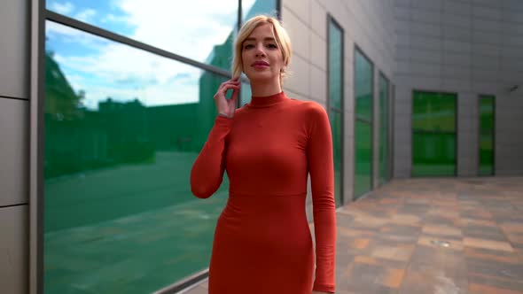 a Blonde in a Red Dress Walks Along a Modern Building and Looks at Her Reflection in the Large Green