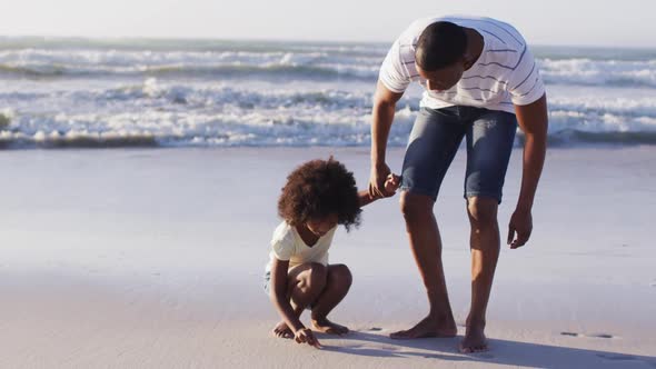 African american father and daughter picking up shells from the sand together at the beach