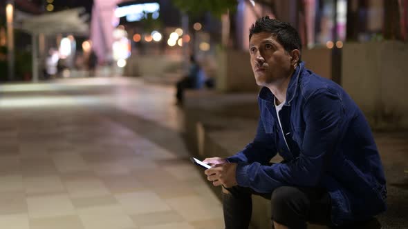 Hispanic Man Thinking and Using Phone While Sitting on the Street in the City at Night