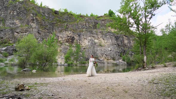 Bride in a White Dress with a Bouquet Walks on the Shore of a Mountain Lake