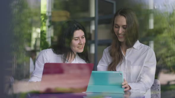 Two Positive Young Women Talking Laughing Surfing Internet on Laptop and Tablet Sitting in Cafe