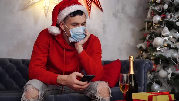 Young Thoughtful Man in Medical Mask and Santa Claus Hat with Smartphone Sitting on Sofa in Room