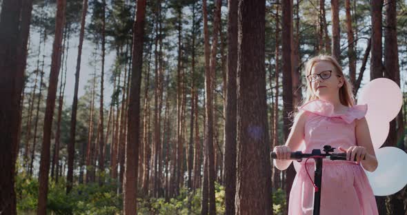 A Girl in a Pink Dress Rides a Scooter in the Park