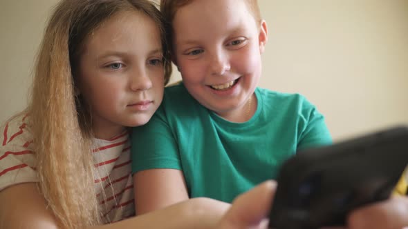 Joyful Small Boy and Girl Watch a Funny Video on Smartphone Sitting on the Sofa at Room