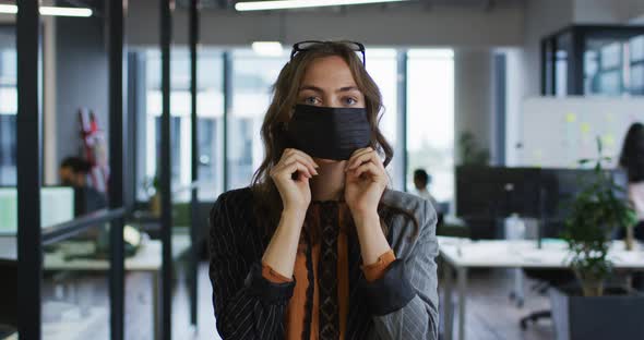 Portrait of caucasian businesswoman adjusting face mask standing in office and looking to camera