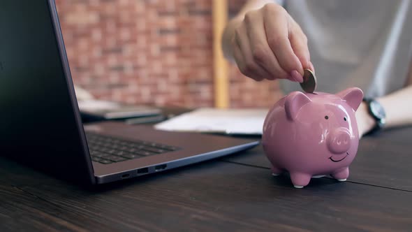 Woman puts coins with hand in the piggy bank, sitting at a desk with a laptop in the office