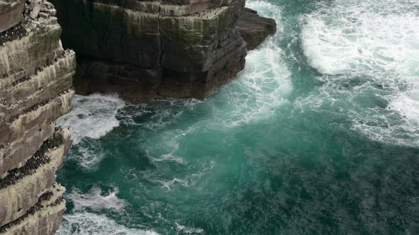 Powerful waves crash against rocks and a sea cliff in the middle of a beautiful, deep teal ocean whi