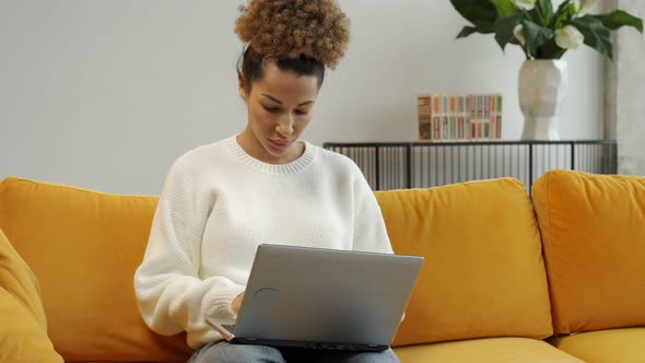 An AfricanAmerican Beautiful Woman Sits on a Yellow Sofa and Uses a Laptop Computer for Work or