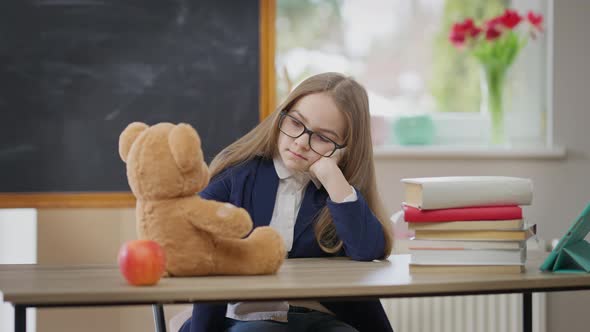 Portrait of Bored Caucasian Schoolgirl with Teddy Bear Sitting in School Classroom Sighing