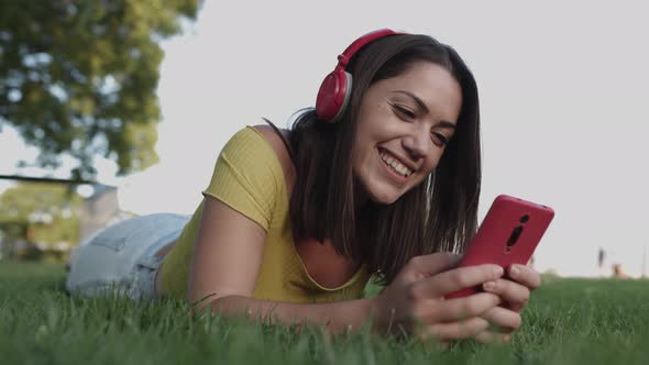 Smiling Young Hispanic Woman Listening to Music with Headphones Lying on Grass