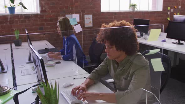 Diverse male and female business colleagues sitting at desks with sneeze guard using computers