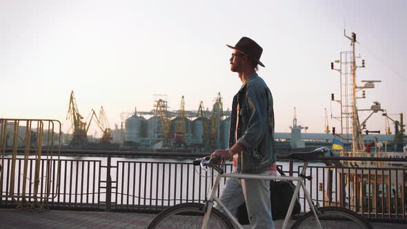 A Young Stylish Hipster with Hat and Glasses Walking with His Bicycle on Sea Port Background During