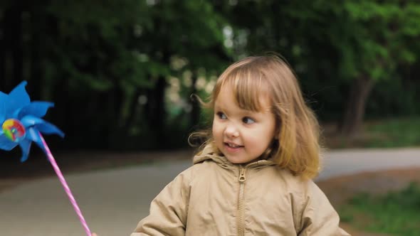 Cute Little Girl Smiles and Looks at Windmill Toy Walks in the Park