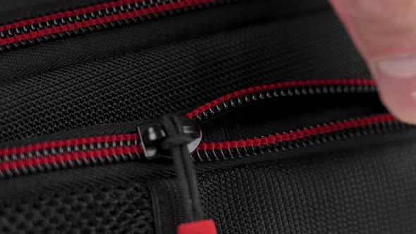 A hand closes the zipper of a black red nylon travel backpack