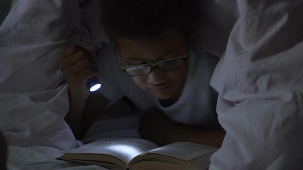 Child Reading Books at Night Under Blanket, Lighting Himself With Flashlight