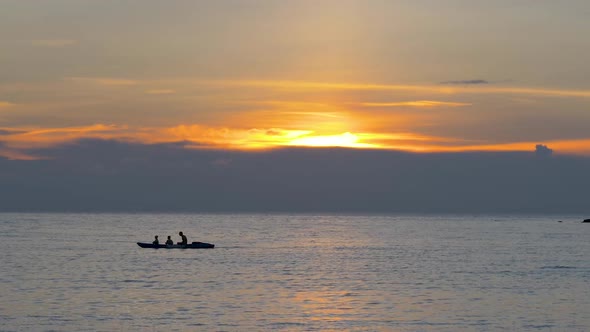 Tourists in a rowboat at colorful orange sunset, silhouettes against the setting sun. ZOOM OUT