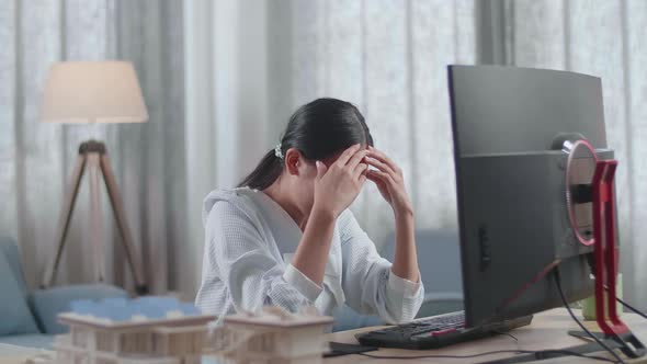 Asian Woman Engineer With The House Model Having A Headache While Working On A Desktop At Home