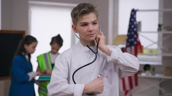 Portrait of Handsome Caucasian Teenage Boy in Medical Gown Using Stethoscope Looking at Camera