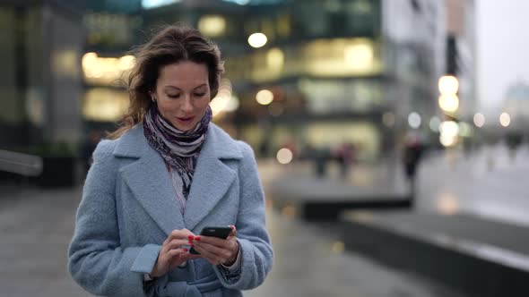 a Woman in a Light Coat and Scarf and with Bright Nails Walks with a Phone in Hands and Smiles at