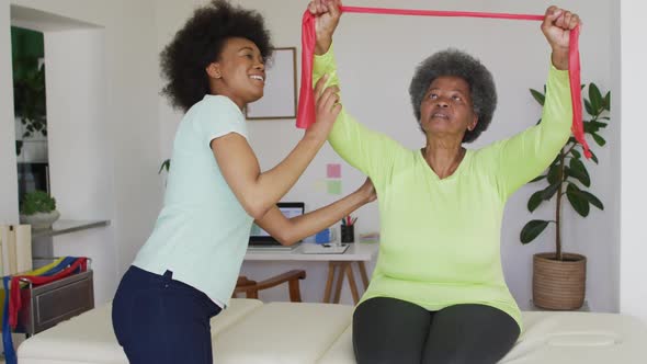 Happy african american female physiotherapist helping senior female patient exercise at home