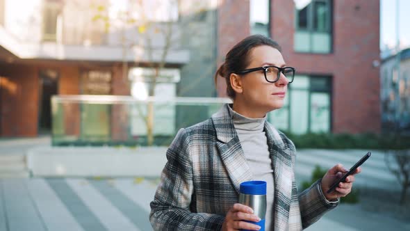 Caucasian Businesswoman with Glasses and in a Coat Walks Through the Business District with Thermo