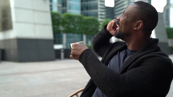 A Cheerful African American Man Spends Time in a Cafe, Sitting on a Coffee Terrace, Drinking a Cup